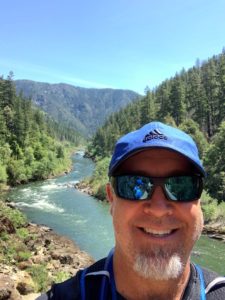 Man with goatee wearing blue had and sunglasses in front of the Rogue River